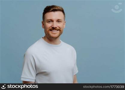 Young positive unshaven guy in white shirt has happy facial expression while looking at camera, handsome hairy man posing against blue studio background. Positive emotions concept. Young positive handsome man in casual wear posing in studio