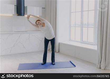 Young positive white woman in sportswear and white t-shirt is stretching at home. Smiling girl is practicing yoga. Concept of healthy lifestyle on quarantine. Morning workout and sport rehabilitation.. Young white woman in sportswear and white t-shirt is stretching at home. Morning workout.