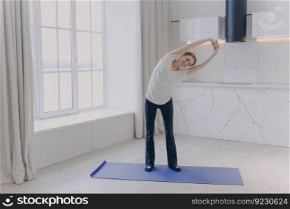Young positive white woman in sportswear and white t-shirt is stretching at home. Smiling girl is practicing yoga. Concept of healthy lifestyle on quarantine. Morning workout and sport rehabilitation.. Young white woman in sportswear and white t-shirt is stretching at home. Morning workout.