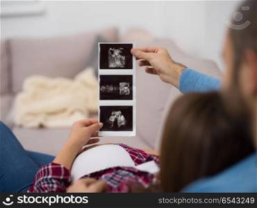 Young pregnant couple looking baby&rsquo;s ultrasound photo while relaxing on sofa at home. pregnant couple looking baby&rsquo;s ultrasound