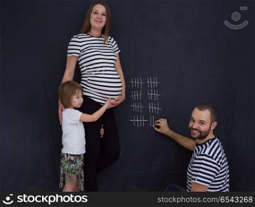 young pregnant couple with cute little daughter accounts week of pregnancy and writing them with chalk on blackboard. happy family accounts week of pregnancy