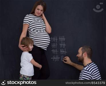 young pregnant couple with cute little daughter accounts week of pregnancy and writing them with chalk on blackboard. happy family accounts week of pregnancy