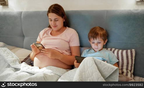 Young pregnant mother with little son lying in bed and using smartphone and tablet computer before going to sleep at night.. Young pregnant mother with little son lying in bed and using smartphone and tablet computer before going to sleep at night