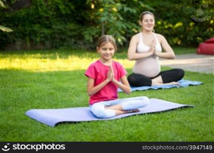 Young pregnant woman and cute girl practicing yoga on grass at park
