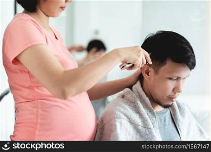 Young Pregnant Woman cutting man hair with clipper during pandemic virus at home. Asian husband getting haircut from his wife during quarantine. Lifestyle of Couple in family barbershop