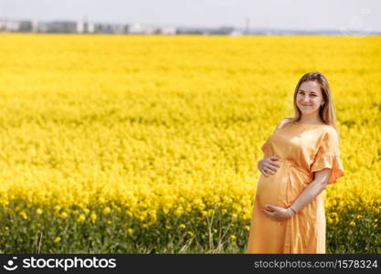 Young pregnant woman in a yellow rapeseed fields holding her belly and wearing yellow dress on sunny summer day.. Young pregnant woman in a yellow rapeseed fields holding her belly and wearing yellow dress on sunny summer day