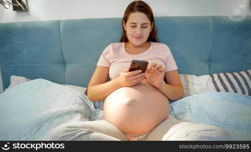 Young pregnant woman in pajamas lying in bed and using smartphone. Pregnant female with phone lying in bed at night.. Young pregnant woman in pajamas lying in bed and using smartphone. Pregnant female with phone lying in bed at night