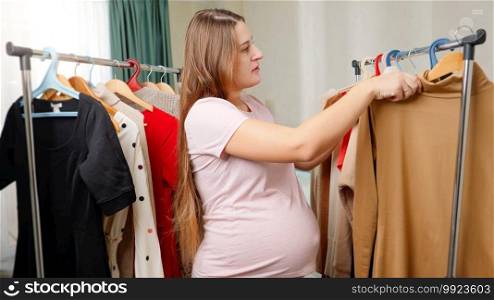 Young pregnant woman in pajamas walking between racks of clothes and searching for something to wear. Pregnant female struggling to find dress that fit.. Young pregnant woman in pajamas walking between racks of clothes and searching for something to wear. Pregnant female struggling to find dress that fit