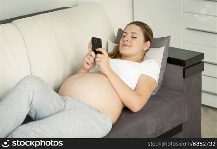 Young pregnant woman lying on sofa and using smartphone