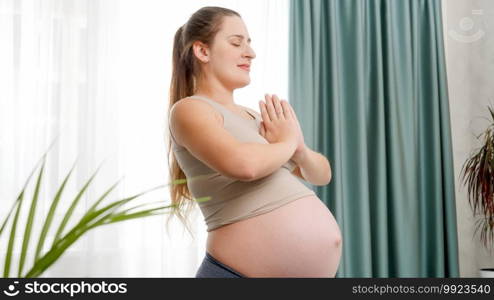 Young pregnant woman meditating and practicing yoga at big window against bright sky. Concept of healthy lifestyle, healthcare and sports during pregnancy.. Young pregnant woman meditating and practicing yoga at big window against bright sky. Concept of healthy lifestyle, healthcare and sports during pregnancy