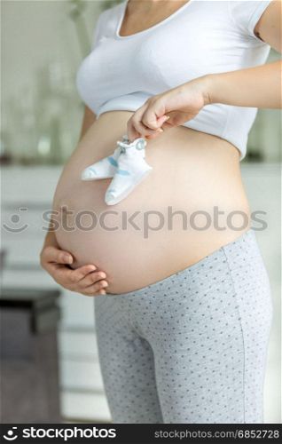 Young pregnant woman posing with baby booties on belly at living room