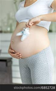 Young pregnant woman posing with baby booties on belly at living room