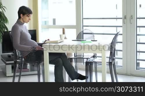 Young pregnant woman reading book for pleasure at home, expectant mother sitting in living room and drinking coffee with mug or tea with cup. People relax, leisure, lifestyle, free time