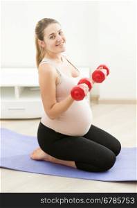 Young pregnant woman sitting on fitness mat and exercising with weights