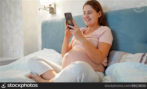 Young pregnant woman talking on video call while sitting on bed at night. Pregnant girl talking to a friend or doctor via video conference on smartphone. Young pregnant woman talking on video call while sitting on bed at night. Pregnant girl talking to a friend or doctor via video conference on smartphone.