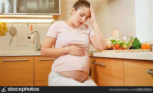 Young pregnant woman with ache in belly resting on chair after cooking and working on kitchen.. Young pregnant woman with ache in belly resting on chair after cooking and working on kitchen