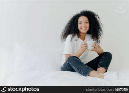 Young pretty African American woman rests in bed at home, drinks hot tea in morning, enjoys domestic atmosphere, tasty drink while relaxes in bedroom. People, leisure, rest and lifestyle concept