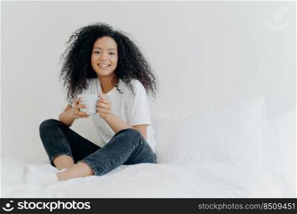 Young pretty African American woman rests in bed at home, drinks hot tea in morning, enjoys domestic atmosphere, tasty drink while relaxes in bedroom. People, leisure, rest and lifestyle concept