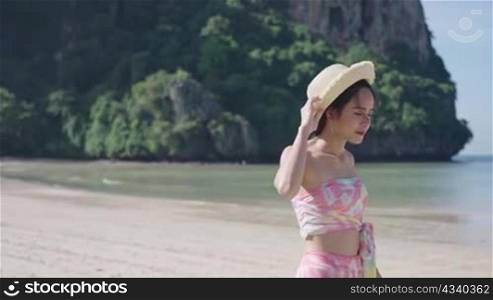 Young pretty asian female walking along Island alone with wind blowing hairs and hat, Travel summer vacation, Asian lady wear straw hat Enjoy summer Holiday, tropical island season hot sunny weather