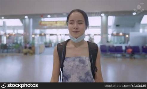 Young pretty asian female wearing protective medical mask walking inside airport terminal, passenger get on escalator during covid19 Corona virus pandemic, backpack traveler distancing concept