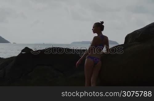 Young pretty blonde female wearing colorful bikini standing between rocks looking into sea and enjoying summer day