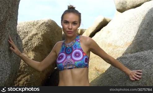 Young pretty blonde female wearing colorful bikini standing between the rocks and looking into the camera