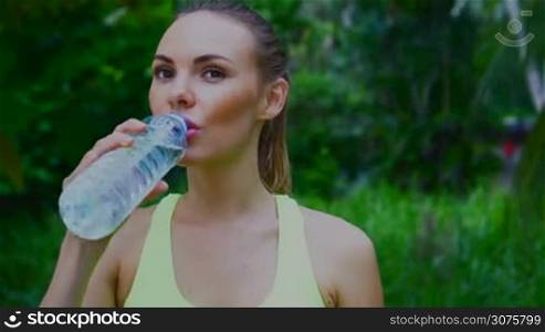 Young pretty blonde female wearing exercise clothing standing in tropical palm trees garden, drinking water after run and enjoying beautiful summer day