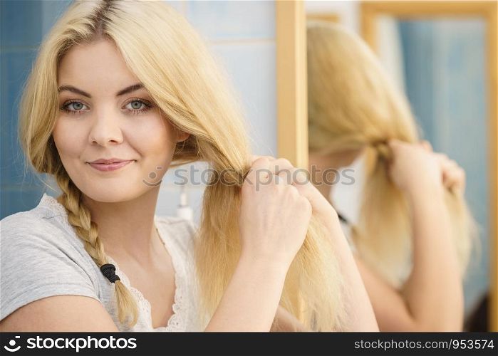 Young pretty blonde woman creating her hairstyle, making braid. Lovely female getting ready. Woman making braid on blonde hair