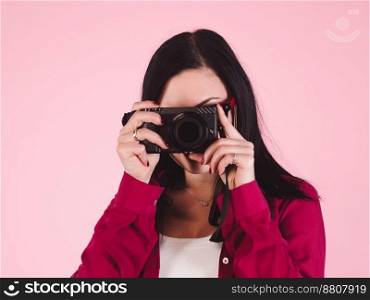 Young pretty brunette woman in pink shirt takes pictures with DSLR camera over colorful background in studio. Girl smiling, flirting and having fun as photographer.