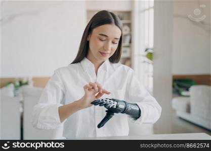 Young pretty disabled girl turns on her bionic prosthetic arm. Female training to use artificial robotic hand after limb loss. People with disabilities, medical high technologies concept.. Young disabled girl turns on her bionic prosthetic arm, training to use artificial robotic hand