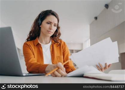 Young pretty european woman in earphones examines reports and charts. Concentrated manager is brainstorming online from home. Corporate internet session on laptop. Remote business concept.. Young pretty european woman examines reports and charts. Manager is brainstorming online from home.