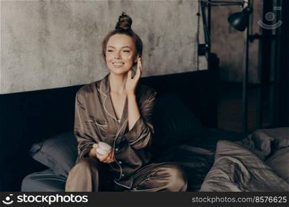 Young pretty european woman in silk pajama takes care of skin and body after waking up in morning at home while applying moisturizing face cream, getting ready for work. Beauty and skincare concept. Young pretty european woman in silk pajamas takes care of her skin and body