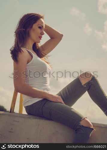 Young pretty fashion model woman sitting on concrete wall wearing white tank top and olive green trousers. Female walking outdoor during warm summer weather.. Fashion model woman outdoor