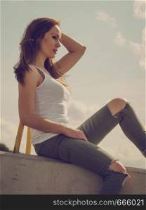 Young pretty fashion model woman sitting on concrete wall wearing white tank top and olive green trousers. Female walking outdoor during warm summer weather.. Fashion model woman outdoor