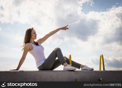 Young pretty fashion model woman sitting on concrete wall wearing white tank top and olive green trousers. Female walking outdoor during warm summer weather.. Fashion model woman outdoor