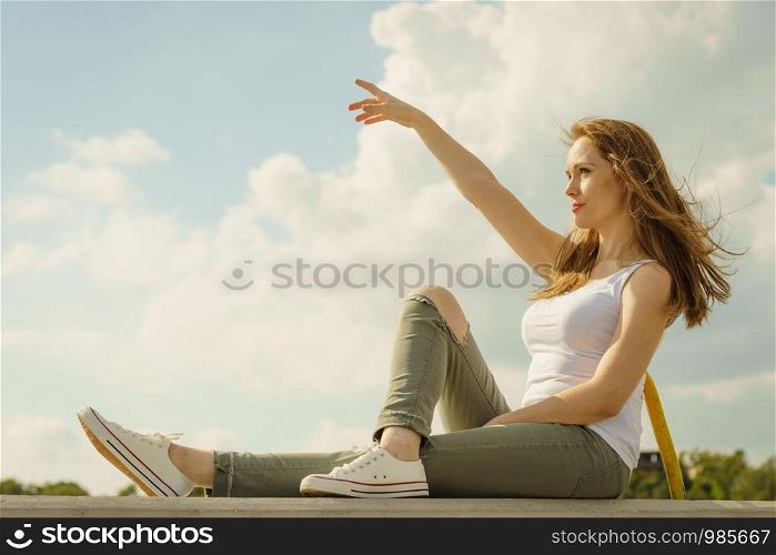 Young pretty fashion model woman sitting on concrete wall wearing white tank top and olive green trousers. Female walking outdoor during warm summer weather.. Fashion model woman outdoor