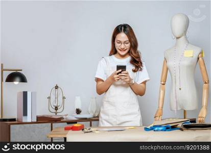 Young pretty female fashion designer stylish used smartphone during working at fashion studio, Tailor woman&rsquo;s taking measurements with measuring tape on mannequin in studio at home