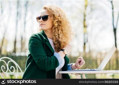 Young pretty female with fluffy blonde hair wearing sunglasses sitting on in the sun in front of laptop translating textes looking around noticing her best friend enjoying sunny weather in the yard