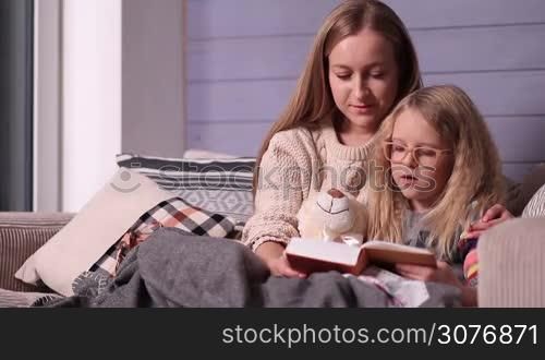Young pretty mother and her little daughter wrapped in a blanket sitting on the sofa in a nice cozy room and reading bedtime story together. Sweet child learning to read with her mother at home.