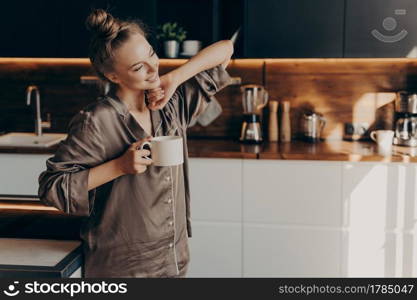 Young pretty relaxed woman in comfy pajama stretching from sleep early in morning while holding cup of coffee in her hand, standing in stylish kitchen interior. Leisure time on average day at home. Young pretty relaxed woman in pajama with cup of coffee in morning