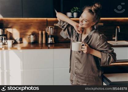 Young pretty relaxed woman in comfy pajama stretching from sleep early in morning while holding cup of coffee in her hand, standing in stylish kitchen interior. Leisure time on average day at home. Young pretty relaxed woman in pajama with cup of coffee in morning