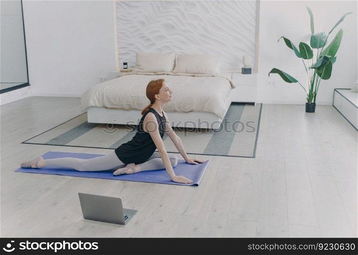 Young pretty white sportswoman practicing yoga on mat in front of computer. Concept of e-learning and home classes. Sport, pilates and stretching asana. Morning routine. Bedroom interior.. Young pretty sportswoman practicing yoga in front of computer. Stretching asana. Morning routine.