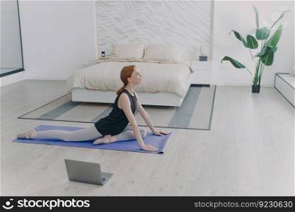 Young pretty white sportswoman practicing yoga on mat in front of computer. Concept of e-learning and home classes. Sport, pilates and stretching asana. Morning routine. Bedroom interior.. Young pretty sportswoman practicing yoga in front of computer. Stretching asana. Morning routine.