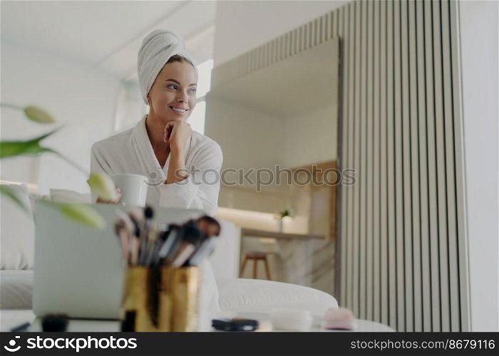Young pretty woman beauty blogger in white bathrobe and hair wrapped in towel looking aside and smiling while relaxing with cup of tea on sofa in living room, managing information in blog on laptop. Female beauty blogger in white bathrobe relaxing on sofa, drinking tea and looking aside