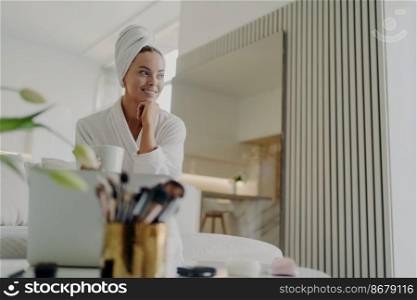 Young pretty woman beauty blogger in white bathrobe and hair wrapped in towel looking aside and smiling while relaxing with cup of tea on sofa in living room, managing information in blog on laptop. Female beauty blogger in white bathrobe relaxing on sofa, drinking tea and looking aside