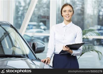 Young pretty woman dealer in showroom standing near car . I will help you to choose car