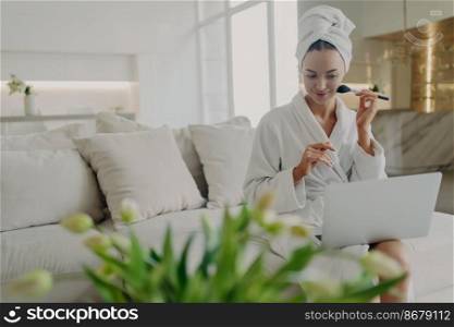 Young pretty woman in bathrobe and towel on head sitting on sofa in modern living room, holding cosmetic brush and doing daily make up while watching video tutorial on laptop, applying powder on face. Young pretty woman in bathrobe doing daily makeup while watching video tutorial on laptop