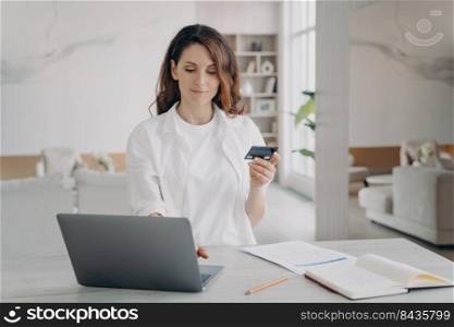 Young pretty woman in white is going to buy through internet. European girl is holding credit card in front of laptop. Booking or purchasing online. E-commerce and financial transaction concept.. Young pretty woman is going to buy through internet. European girl is holding credit card.