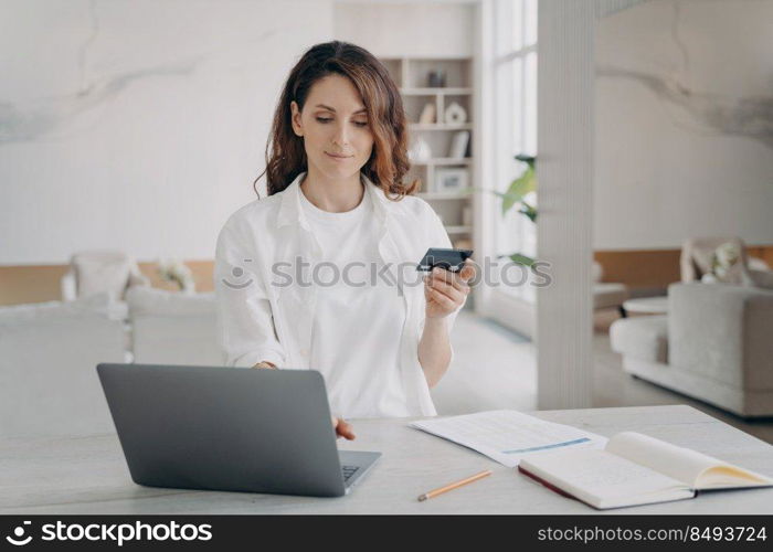 Young pretty woman in white is going to buy through internet. European girl is holding credit card in front of laptop. Booking or purchasing online. E-commerce and financial transaction concept.. Young pretty woman is going to buy through internet. European girl is holding credit card.