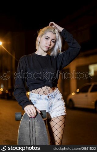 Young pretty woman looking camera in shorts while standing on the street holding a skateboard at night in the city